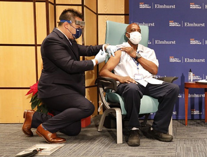 NYC Health + Hospitals health worker, wearing mask and suit, kneels and administers vaccine to a patient at a vaccnination event