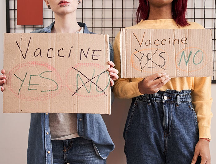 stock photo of two people holding signs saying Vaccine Yes No - one has yes crossed out, one has no crossed out